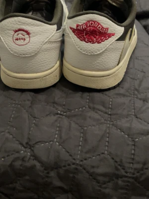 Nike Air Jordan 1 Low Travis Scott - Säljer ett par Nike Air Jordan 1 Low Travis Scott med vit och olivgrön ovandel, röda detaljer och klassisk Air Jordan Wings-logga på hälen. Skorna har en unik broderad smiley på ena hälen och är tillverkade i läder med textilfoder. Perfekta för dig som gillar streetwear.