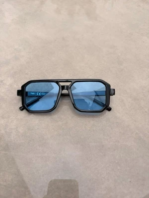 Svarta solglasögon med blå glas - Snygga solglasögon med svart, kantig båge och blåtonade glas. Glasögonen har en modern, geometrisk form och passar perfekt för dig som vill sticka ut. Tillverkade i syntetmaterial för en lätt känsla.❤️🕶️⛱️