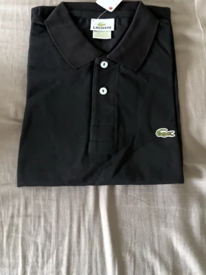 Svart pikétröja från Lacoste - Klassisk svart pikétröja från Lacoste med vit knappstängning och den ikoniska krokodilloggan på bröstet. Tröjan har krage och korta ärmar, tillverkad i mjuk bomull som känns skön mot huden. Perfekt till jeans eller shorts för en clean look.