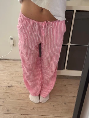Rosa randiga byxor med dragsko - Säljer ett par rosa byxor med vita vertikala ränder och dragsko i midjan. Byxorna har en avslappnad passform och är perfekta för en chill look. Materialet ser ut att vara mjukt och luftigt, troligen bomull eller linne. Perfekta för dig som gillar bekväma och snygga plagg.