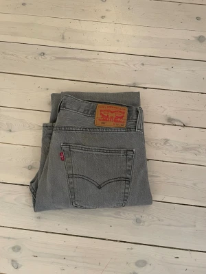 Grå Levi's 501 raka jeans - Säljer ett par grå Levi's 501 jeans med rak passform och klassisk design. Jeansen har den ikoniska läderlappen bak i midjan och röd Levi's-tagg på bakfickan. Tillverkade i kraftig denim, perfekta för en avslappnad och trendig look.
