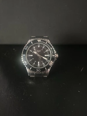 Svart och silver klocka från Regal - Stilren klocka från Regal med svart urtavla och silverfärgade detaljer. Klockan har en klassisk rund form, tydliga siffror och markeringar samt en robust länk i metall. Perfekt accessoar för dig som gillar en clean och tidlös look. Säljs billigare då batteriet är slut!
