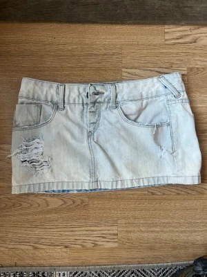 Ljus jeanskjol med slitningar - Säljer en kort ljusblå jeanskjol med slitna detaljer och råa kanter. Kjolen har klassiska fickor fram och bak samt bälteshällor. Perfekt för dig som gillar en avslappnad och trendig stil.