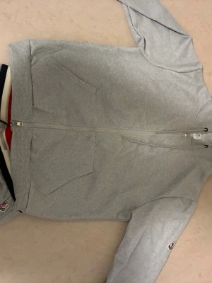 Ljusgrå hoodie från Moncler - Snygg ljusgrå hoodie från Moncler med dragkedja framtill och två stora fickor. Klassisk Moncler-logga på ärmen och mjukt bomullsmaterial. Perfekt för en avslappnad och trendig look.