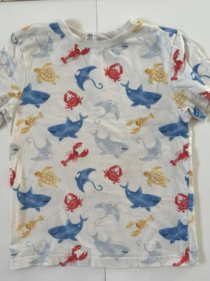 Vit t-shirt med havsdjur strl 122 - Cool vit t-shirt med färgglatt havsdjursmönster, inklusive blåa och gråa hajar, röda krabbor, gula sköldpaddor och mantor.     Små fläckar finns, se bild