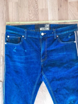 Blå jeans från Love Moschino - Snygga blå jeans från Love Moschino med klassisk femficksmodell och kontrastsömmar. Jeansen har en cool tvättad look och vita detaljer längs sidorna. Tillverkade i slitstarkt denimtyg, perfekta för dig som gillar streetwear och vill sticka ut.