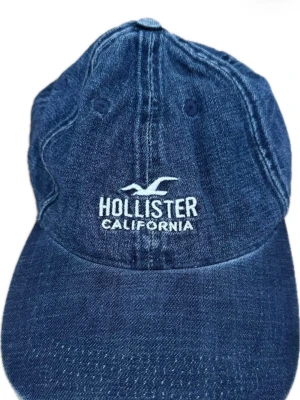 Mörkblå denimkeps från Hollister - Snygg mörkblå keps i denim från Hollister med broderad vit logga och texten 'California' framtill. Klassisk böjd skärm och justerbar passform baktill☀️