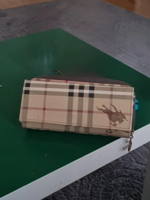 Burberry Rutig plånbok med hästmotiv - Snygg plånbok i beige och brunt med klassiskt rutigt mönster och hästlogga. Plånboken har flera kortfack, fönster för ID och tryckknappsstängning. Utsidan är i tåligt konstläder och insidan i brunt material.