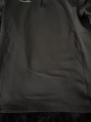 Svart träningströja från Under Armour - Svart långärmad träningströja från Under Armour med halv dragkedja framtill och logga på bröstet. Perfekt för löpning eller gym. Materialet är mjukt och stretchigt för maximal rörelsefrihet.