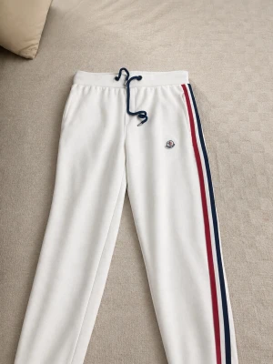 Vita mjukisbyxor från Moncler - Snygga vita mjukisbyxor från Moncler med blå och röd sidrand längs ena benet och broderad Moncler-logga på låret. Byxorna har dragsko i midjan och är tillverkade i mjukt material för maximal komfort. Perfekta för en chill och sportig look.