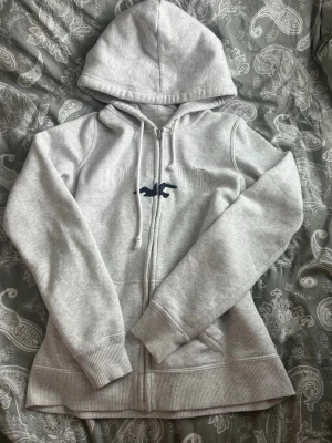 Ljusgrå zip hoodie från Hollister - Ljusgrå hoodie med dragkedja från Hollister. Klassisk modell med huva och dragsko, ribbade muddar och nederkant. Broderad Hollister-logga och fågel på bröstet i mörkblått. Mjuk bomullsblandning som känns skön mot huden.