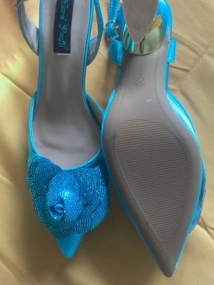 Blå glittriga pumps med blomdetalj - Snygga blå pumps med spetsig tå och öppen häl, dekorerade med en stor glittrig blomma framtill. Skorna har en elegant rem runt hälen och är tillverkade i ett glansigt material som ger en festlig känsla. Perfekta för dig som vill sticka ut med färg och detaljer.