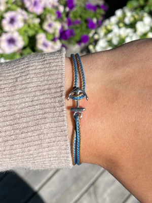 Blått och brunt armband med ankare - Säljer ett snyggt armband med dubbla snören i blått och brunt. Armbandet har ett coolt ankare i silverfärgad metall som lås. Perfekt för dig som gillar marina detaljer och stilrena accessoarer.
