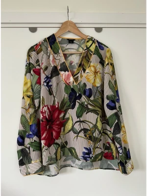 Lindex - Blommig & randig blus med v-ringning - storlek 38 - Härlig blus med långa ärmar och ett färgglatt blommönster från Lindex i storlek 38. Klassisk V-ringning med en dekorativ gul kant som även återfinns vid ärmslut och nederkant. Mönstret inkluderar ränder i brunt och vitt som bakgrund till de stora blommorna i rött, gult, rosa och blått, samt gröna blad. Otroligt härlig blus som passar till både vardag och fest. Mycket gott skick, som ny!