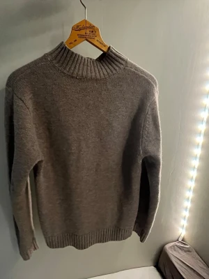 Grå stickad tröja med hög krage - En enkel och stilren grå stickad tröja med hög ribbad krage och långa ärmar. Tröjan har ribbstickade muddar vid ärmslut och nederkant. Perfekt för att hålla sig varm under kyliga dagar.