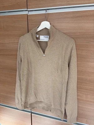 Beige halvzip tröja Selected Homme - Snygg beige tröja från Selected Homme med halvzip och hög krage. Tillverkad i mjukt material, perfekt för lager på lager. Långärmad modell med ribbade muddar vid ärmslut och nederkant. Enkel och stilren design som passar till mycket.