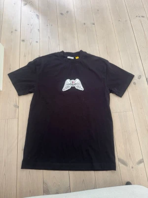 Svart t-shirt med ving- och hjärtdetalj - Säljer en svart t-shirt med korta ärmar och ett broderat motiv av vita vingar och ett rött hjärta på bröstet. T-shirten har en klassisk passform och är tillverkad i mjuk bomull. Perfekt för dig som gillar unika detaljer.