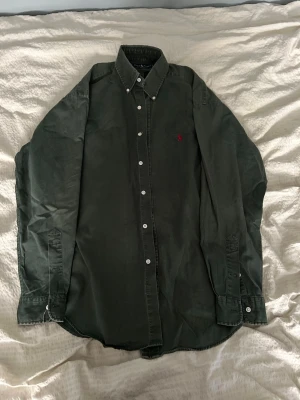 Mörkgrön skjorta från Ralph Lauren - Klassisk mörkgrön skjorta från Ralph Lauren i modellen Blaire. Skjortan är tillverkad i 100% bomull och har vita knappar framtill samt en liten röd broderad logga på bröstet. Perfekt för en clean och stilren look. Hör av er vid frågor!😃