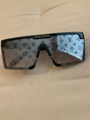 Lv solglasögon  - Snygga solglasögon med svart båge och stora, rektangulära glas. Glasen har ett grått LV-monogrammönster som ger en exklusiv vibe. Märkeslogga i guld på bågen och över glaset. Perfekta för dig som vill sticka ut med lyxig stil.