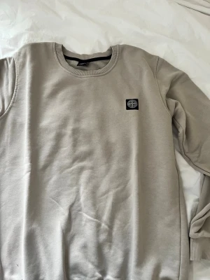 Beige sweatshirt från Stone Island - Snygg beige sweatshirt från Stone Island med rund halsringning och diskret logotyp-patch på bröstet. Tröjan har en klassisk passform och är tillverkad i mjukt bomullsmaterial. Perfekt för en clean och stilren look.