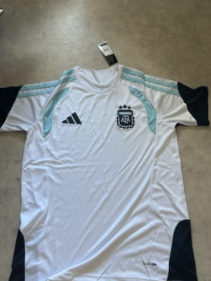 Argentina Adidas fotbollströja vit - Snygg vit fotbollströja från Adidas med Argentinas landslagsemblem och tre stjärnor på bröstet. Tröjan har ljusblå detaljer på axlarna, svarta inslag på ärmar och sidor samt Climacool-material som andas. Perfekt för match eller träning.