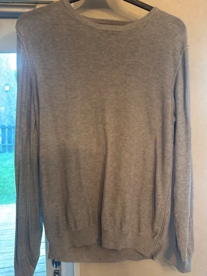 Ljusgrå tröja från Bläck - En stilren ljusgrå tröja från Bläck med rund halsringning och långa ärmar. Tröjan är tillverkad i ett mjukt, tunt material och har ribbade muddar vid ärmslut och nederkant. Perfekt för lager på lager och enkel att matcha med olika outfits.