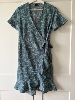 Prickig omlottklänning Vero Moda - Supersöt blå/grön omlottklänning från Vero Moda med vita prickar, volangkant nertill och knyt i midjan. Klänningen har korta ärmar och v-ringning, perfekt för en fräsch och trendig look. Använt en gång.