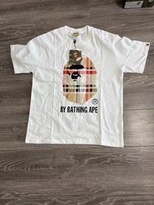 Vit BAPE t-shirt med tryck - Säljer en vit t-shirt från BAPE med stort ikoniskt ap-tryck på bröstet i rutigt och kamouflagemönster. T-shirten har korta ärmar och rund hals. Klassisk streetwear-stil med 'BY BATHING APE' text framtill.