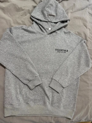 Grå Essentials Fear of God hoodie - Snygg grå hoodie från Essentials Fear of God med svart logga på bröstet. Klassisk passform med huva, ribbade muddar och magficka. Tillverkad i mjukt bomullsmaterial som känns skönt mot huden. Perfekt för en avslappnad och trendig look.