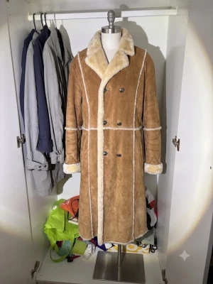 Lång brun fårskinnskappa retro - Snygg lång brun fårskinnskappa med vit fuskpäls på krage, ärmslut och insida. Dubbelknäppt med stora knappar och markerade sömmar. Perfekt för kalla dagar och ger en retrovibe till din outfit.