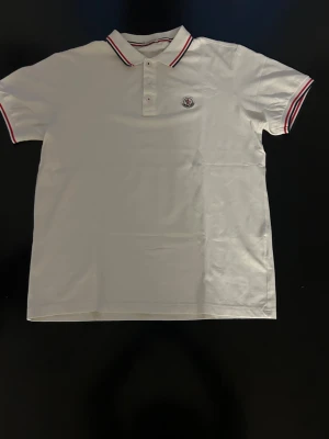 Vit Moncler piké med rödblå detaljer - Klassisk vit pikétröja från Moncler med korta ärmar och krage. Tröjan har rödblå ränder på krage och ärmslut samt Moncler-logga på bröstet. Tillverkad i mjuk bomull för en bekväm känsla och stilren look.