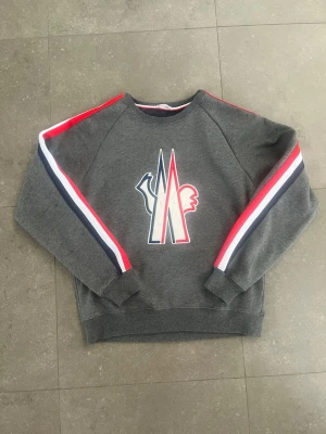 Grå sweatshirt från Moncler - Snygg grå sweatshirt från Moncler med stort logotryck på bröstet och ränder i rött, vitt och blått längs ärmarna. Utmärkt skick, storlek M