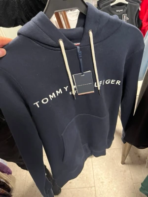 Marinblå hoodie från Tommy Hilfiger - Snygg marinblå hoodie från Tommy Hilfiger med vit logga över bröstet och vita dragsnören i huvan. Klassisk känguruficka framtill och mjukt material som känns skönt mot huden. Perfekt för en avslappnad och trendig stil.