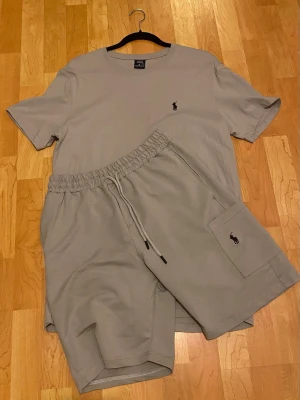 Beige set från Polo Ralph Lauren - Snyggt beige set från Polo Ralph Lauren med t-shirt och shorts. Båda plaggen har den klassiska broderade loggan i svart. T-shirten har rund hals och kort ärm, shortsen har resårmidja med dragsko och en ficka på sidan. Perfekt för en avslappnad och stilren look.