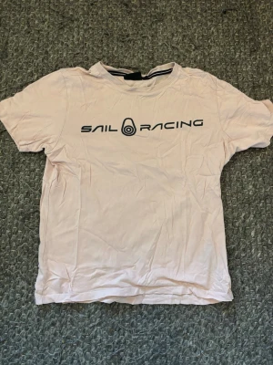 Ljusrosa Sail Racing t-shirt - Säljer en ljusrosa t-shirt från Sail Racing med svart logga tryckt på bröstet. Klassisk rund halsringning och korta ärmar. Perfekt för dig som gillar enkel och stilren design. Materialet är mjuk bomull som känns skönt mot huden.