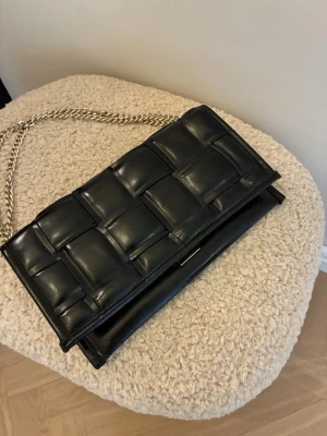 Svart kuvertväska Steve Madden - Snygg svart kuvertväska från Steve Madden med flätad framsida och guldfärgad kedjerem. Väskan har en stilren, rektangulär form. Perfekt accessoar för att lyfta din outfit.