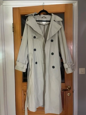Ljusbeige trenchcoat med bälte - Stilren ljusbeige trenchcoat med dubbelknäppning och stora svarta knappar från H&M i superbra kvalitet. Klassisk krage, bälte i midjan och justerbara ärmslut. Två sidofickor och slits bak. Perfekt för lager-på-lager och en tidlös look. Säljer då den är för lång för mig
