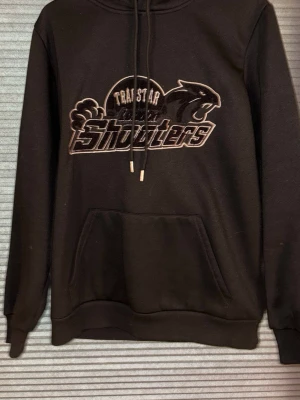 Svart Trapstar hoodie med tryck - Säljer en svart hoodie från Trapstar med stort broderat tryck på bröstet där det står 'Trapstar Shooters' och en panter. Hoodien har huva med dragsko och en stor magficka. Perfekt för dig som gillar streetwear och vill sticka ut.