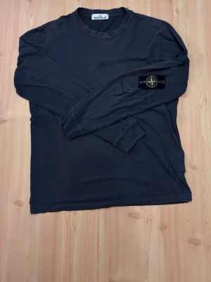 Grå långärmad tröja Stone Island - Grå långärmad tröja från Stone Island med klassisk rund hals och ikonisk patch med logga på vänster ärm. Tillverkad i mjuk bomull för en skön och avslappnad look. Perfekt till streetwear och enkel att matcha med andra plagg. Tröjan är äkta.