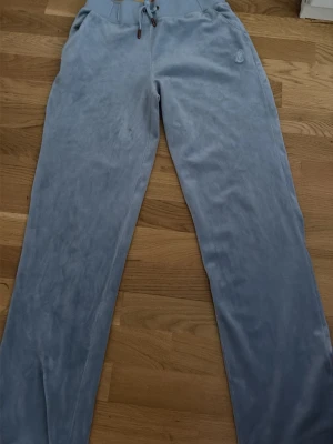 Blå velour mjukisbyxor från Juicy Couture - Säljer ett par ljusblå mjukisbyxor i mjuk velour från Juicy Couture. Byxorna har raka ben, dragsko i midjan och broderad JC-logga på ena sidan. Perfekta för en chill och avslappnad stil äkta 