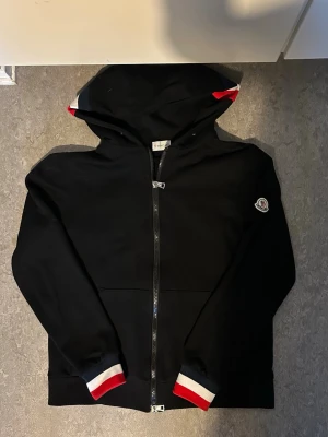 Svart Moncler hoodie med röd/vita detaljer - Svart hoodie från Moncler med dragkedja framtill och huva. Ärmsluten och huvan har snygga rödvita ränder. Moncler-logga på vänster ärm. Perfekt för dig som vill ha en stilren men ändå sportig look.