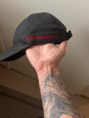 Svart keps från Gucci med GG-mönster - Snygg svart keps från Gucci med klassiskt GG-monogram över hela tyget och en grön-röd rand på sidan. Kepsen har böjd skärm och justerbar rem bak. Tillverkad i ett slitstarkt tyg som känns lyxigt och trendigt.