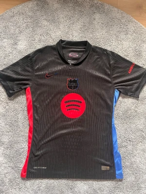 Svart Nike FC Barcelona t-shirt - Säljer en svart FC Barcelona t-shirt från Nike med röd och blå detalj på sidorna. Tröjan har korta ärmar, klubbmärke och Spotify-logga på bröstet samt UNHCR-tryck på ryggen. Tillverkad i lätt och ventilerande polyester, perfekt för fotbollsfans. Tröjan har inga tecken på användning. Perfekt för dig med bra stil🤩