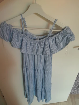Blåvit randig offshoulder klänning - Söt blåvit randig klänning med volang upptill och smala axelband. Offshoulder-modell som ger en somrig känsla. Lätt och luftig, perfekt för varma dagar. Klänningen har en avslappnad passform och faller fint.