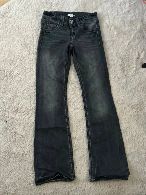 Svarta/mörkgråa bootcut jeans med slitningar - Säljer ett par svarta/mörkgråa bootcut jeans med snygga slitningar och kontrastsömmar. Jeansen har fem fickor, dubbla knappar i midjan och klassisk dragkedja. Perfekta för en avslappnad och trendig look. Från Gina Young, storlek 164. Midjemått rakt över är 34,5cm, de är 20cm ”höga” i midjan och innerbenslängden är 79cm.