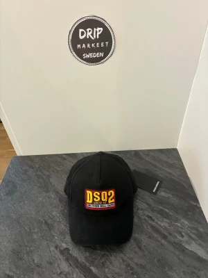 Svart DSQ2 keps mee patch - Snygg svart keps från DSQUARED2 med broderad patch framtill där det står 'DSQ2 SINCE 64 BROTHER HILL GANG' i gult, vitt och rött. Klassisk böjd skärm och justerbar rem bak. Perfekt för dig som vill ha en streetwear vibe.