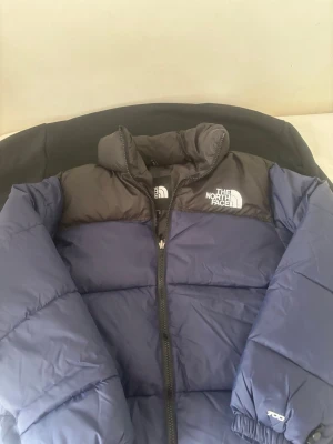 Blå och svart pufferjacka The North Face - Snygg blå och svart pufferjacka från The North Face med hög krage och dragkedja framtill. Jackan har logga på bröstet och ärmen, samt quiltad design och tjock vaddering som håller dig varm. Perfekt för kalla vinterdagar och riktigt trendig streetstil.