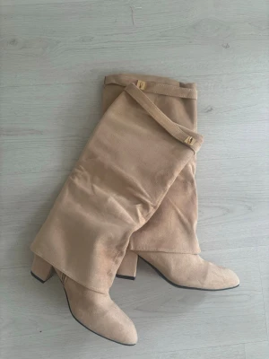Beiga overknee stövlar med klack - Snygga beiga overknee stövlar med bred klack och spetsig tå. Tillverkade i mockaimitation med dekorativa spännen upptill. Perfekta för att lyfta din outfit med en trendig touch.