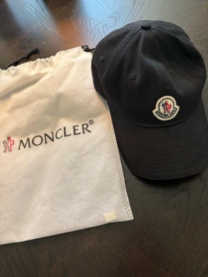 Svart keps från Moncler med logga - Säljer en svart keps från Moncler med det klassiska broderade logotypmärket framtill. Kepsen har böjd skärm och justerbar rem bak. Tillverkad i bomullstyg och levereras med Moncler dustbag.