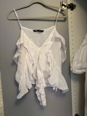 Vit volangtop från Zara - Säljer en superfin vit topp från Zara med tunna axelband och v-ringning. Toppen har volangdetaljer framtill och är tillverkad i ett lätt och luftigt material, perfekt för varma dagar. Snygg att styla med jeans eller kjol! Är lite skrynklig på bilderna, men enkelt att bara stryka
